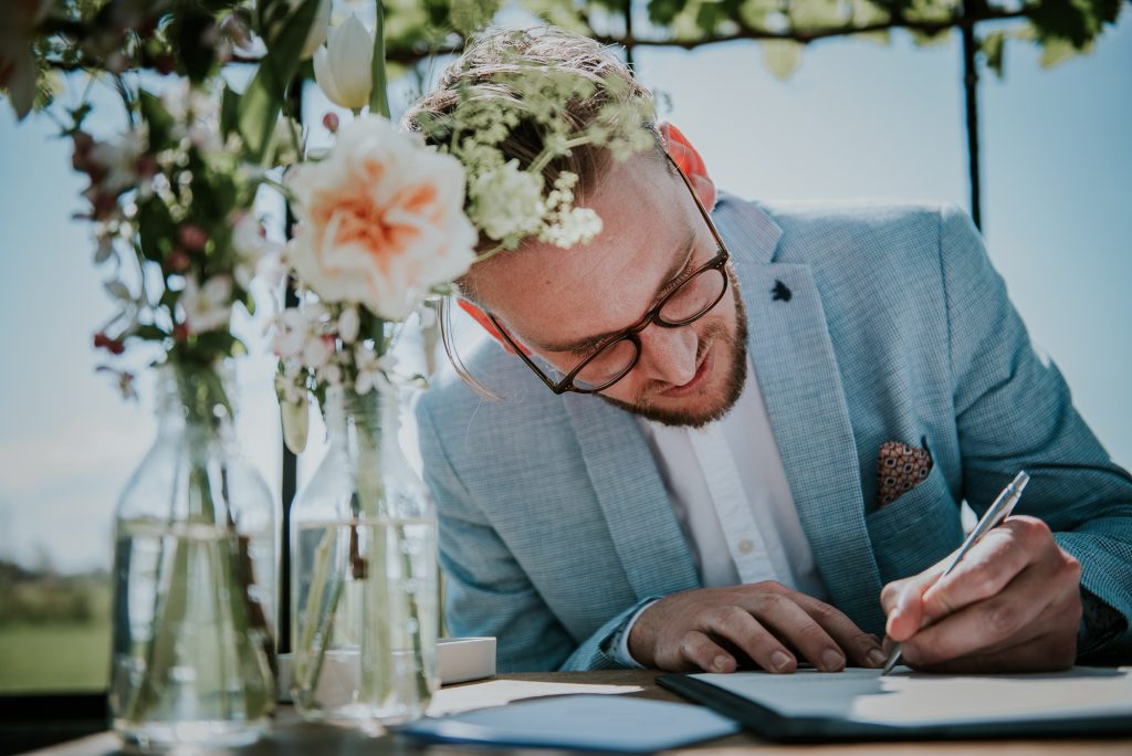 Getuige ondertekent de huwelijksakte tijdens de trouwceremonie in de kas van it Flinkeboskje. Bruidsreportage door bruidsfotograaf Nickie Fotografie uit Dokkum, Friesland