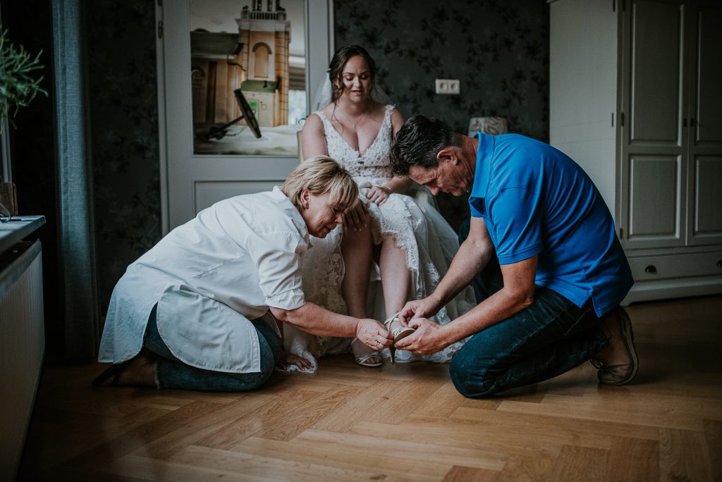 Vader en moeder van de bruid helpen allebei met het aandoen van de bruidsschoenen. Bruidsreportage door bruidsfotograaf NIckie Fotografie uit Dokkum, Friesland.