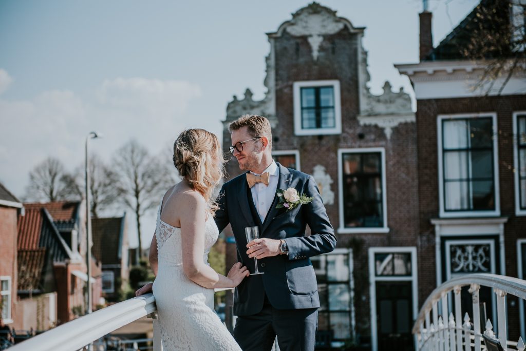 Trouwen in Dokkum, Friesland. Trouwfotografie door trouwfotograaf Nickie Fotografie. Bruidegom,met glas champagne , en bruid op het witte bruggetje met prachtige oude gevels op de achtergrond.