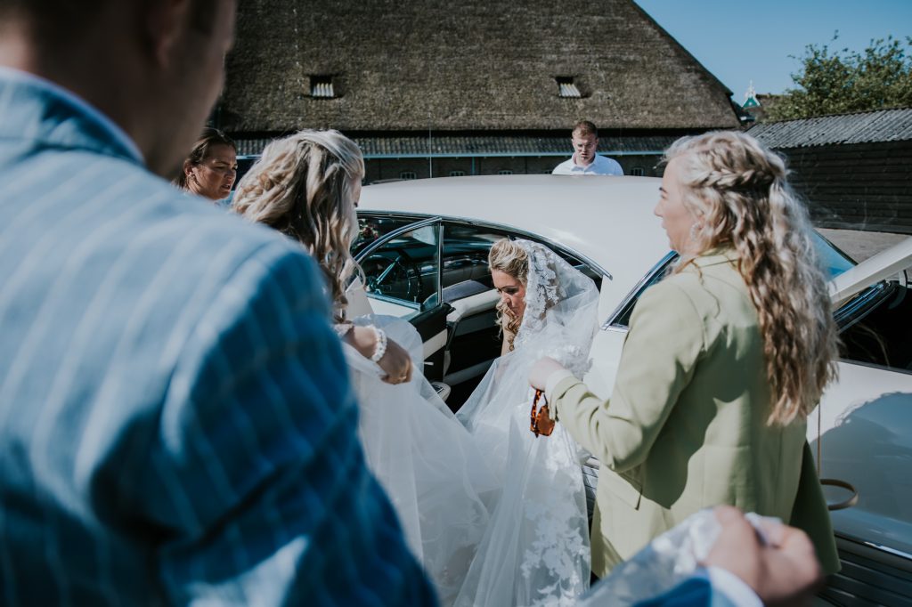 Bruid wordt geholpen bij het instappen in de bruidsauto. Huwelijksreportage door huwelijksfotograaf NIckie Fotografie uit Dokkum.