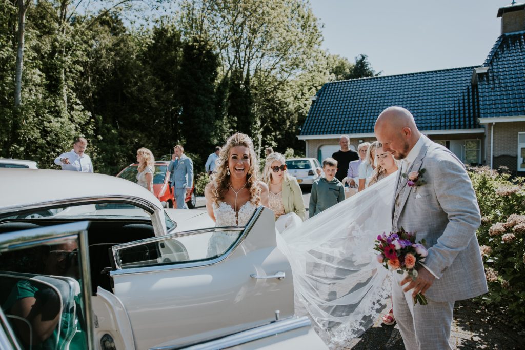 Bruidegom helpt zijn bruid bij het instappen van de prachtige oude witte Cadillac. Huwelijksreportage door huwelijksfotograaf Nickie Fotografie.