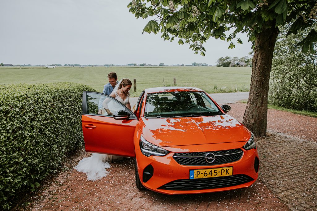 Het bruidspaar stapt in de oranje bruidsauto. Trouwfotografie door trouwfotograaf Nickie Fotografie uit Dokkum, Friesland.