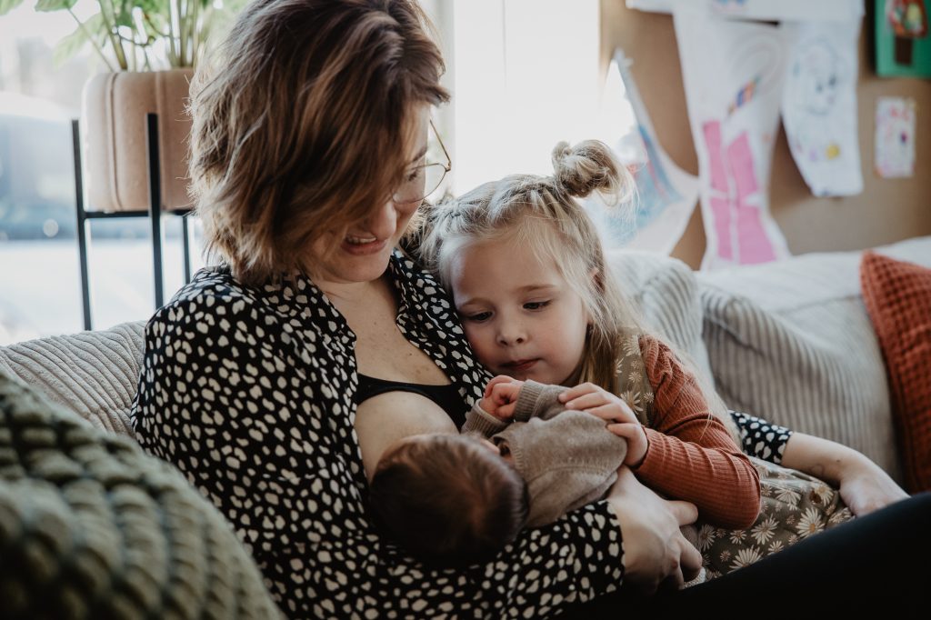 Lifestyle babyfotografie Friesland, Eastermar. Moeder geeft borstvoeding terwijl de grote zus nieuwschierig aan het kijken is. Fotoreportage door fotograaf Nickie Fotografie.
