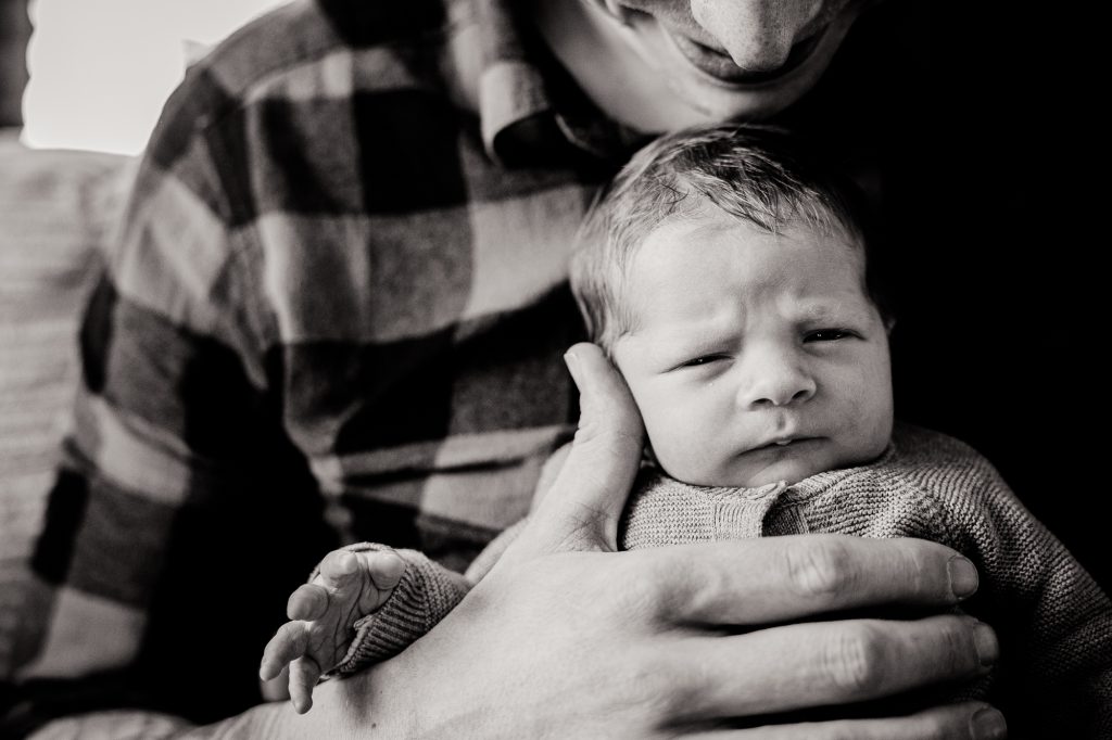 Zwart-wit newbornshoot door babyfotograaf Nickie Fotografie uit Dokkum, Friesland. Portret van baby op schoot bij zijn vader.