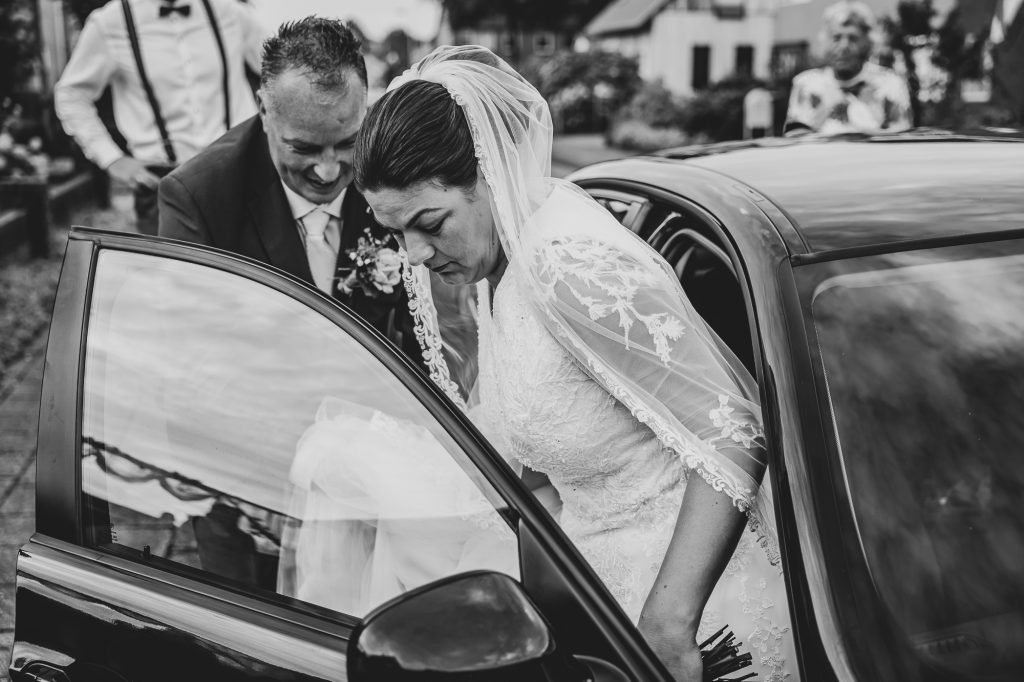 Trouwfotograaf Friesland, Nickie Fotografie. Bruid stapt in de trouwauto en bruidegom helpt hierbij met haar sleep.