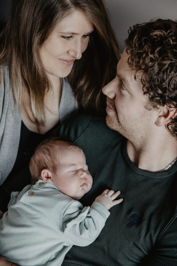 Newbornshoot Dokkum door newbornfotograaf Nickie Fotografie. Lifestyle foto van moeder en vader met de kleine. Vader heeft de baby tegen zijn borst aan.