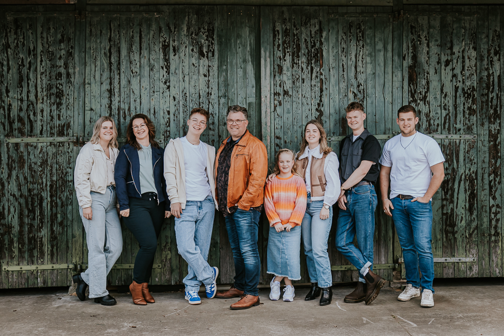 Fotograaf Friesland. Gezinsshoot Kollumerzwaag. Groepsportret van het gezin met aanhang voor oude houten verweerde groene garage deur door fotograaf Nickie Fotografie.