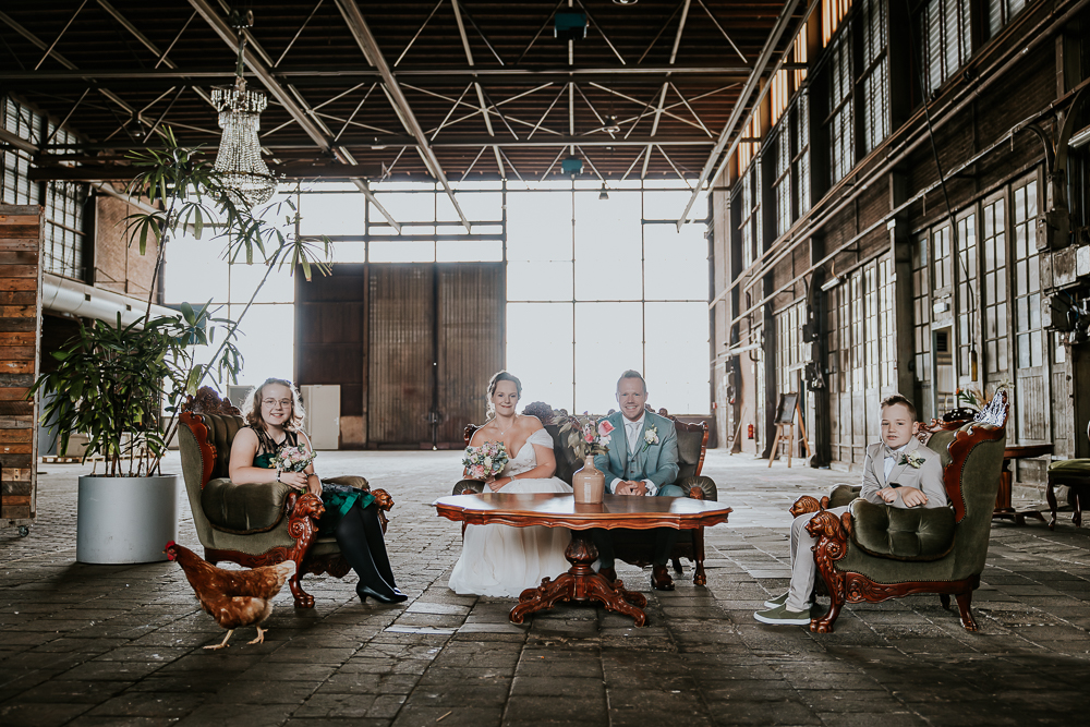 Fotoshoot van het bruidspaar met hun kinderen in de Sluisfabriek in Drachten. Het gezin zit op de antieke stoeltjes in de grote hal. Bruidsfotografie door trouwfotograaf Nickie Fotografie uit Friesland.