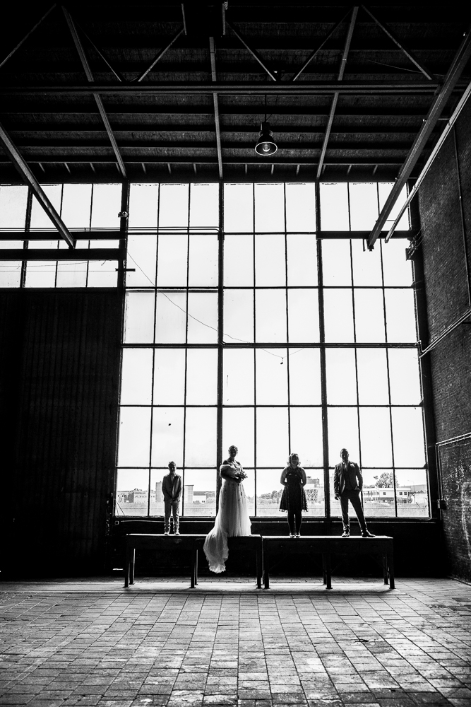 Fotoshoot Sluisfabriek Drachten. Zwart-wit gezinsportret van het bruidspaar, met tegenlicht voor de grote ramen in de grote fabriekshal. Bruidsfotografie door trouwfotograaf Nickie Fotografie.