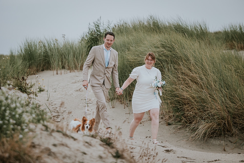 Trouwen op Ameland. Het bruidspaar wandelt met hun hond door de duinen. Bruidsfotografie door bruidsfotograaf Nickie Fotografie.