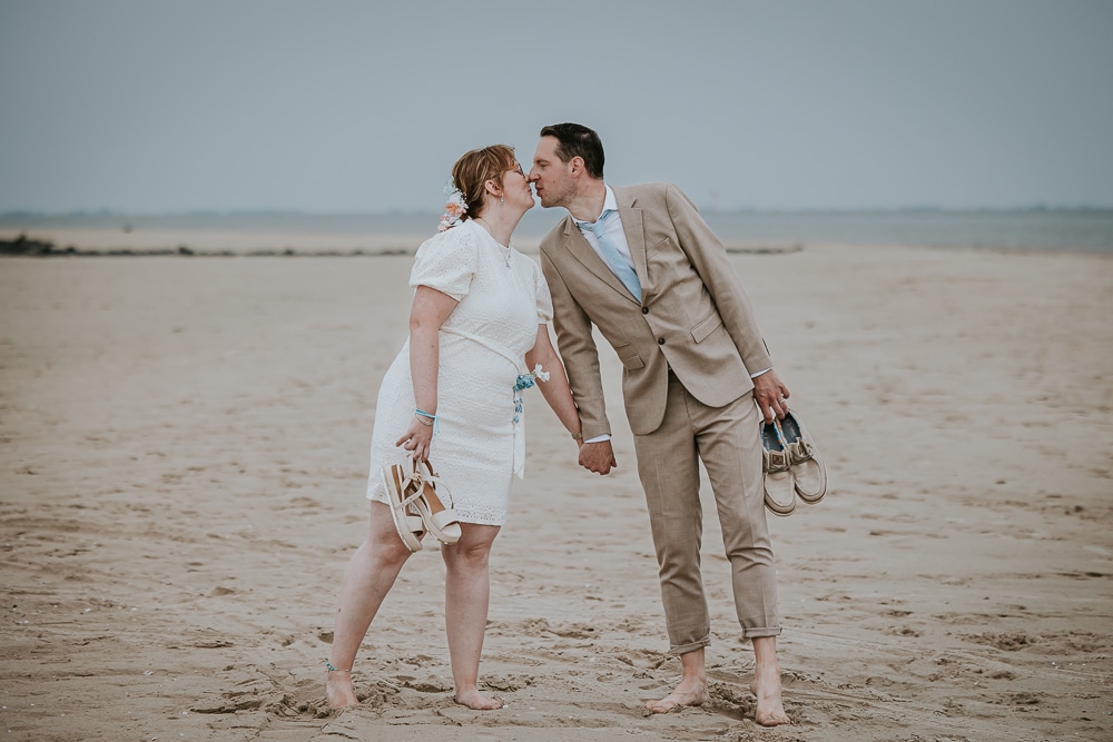 Trouwen op Ameland. Fotoshoot van het bruidspaar, op blote voeten, op het strand. Bruidspaar met de trouwschoenen in de hand. Huwelijksreportage door huwelijksfotograaf Nickie Fotografie uit Friesland.