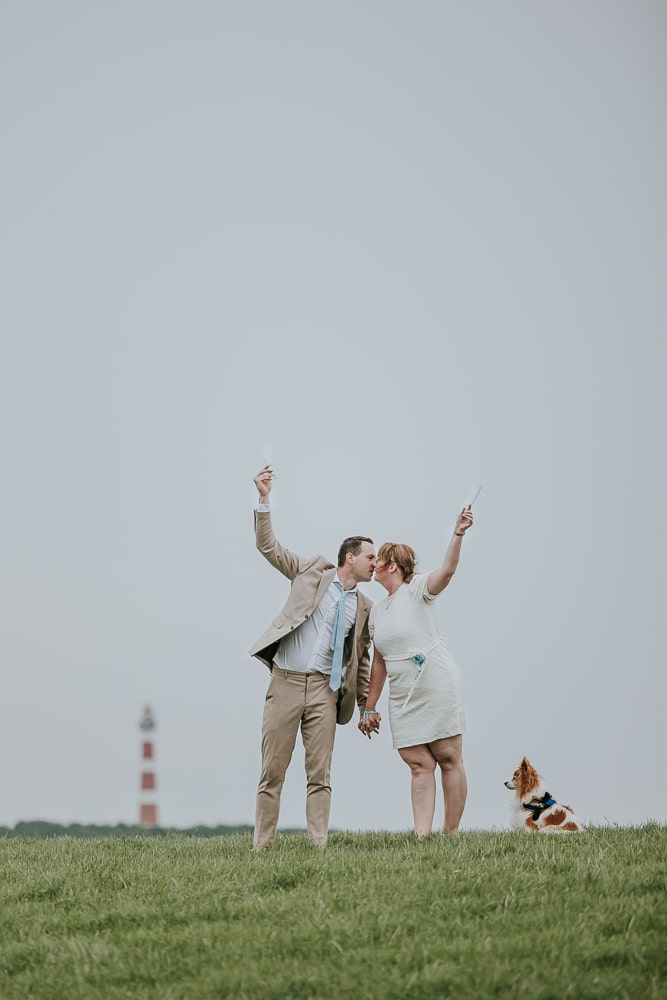 Trouwen op Ameland. Trouwfoto van het bruidspaar en hun hond met de champagne glazen in de lucht met op de achtergrond de roood-wit gestreepte vuurtoren. Huwelijksfotografie door trouwfotograaf Nickie Fotografie uit Friesland.