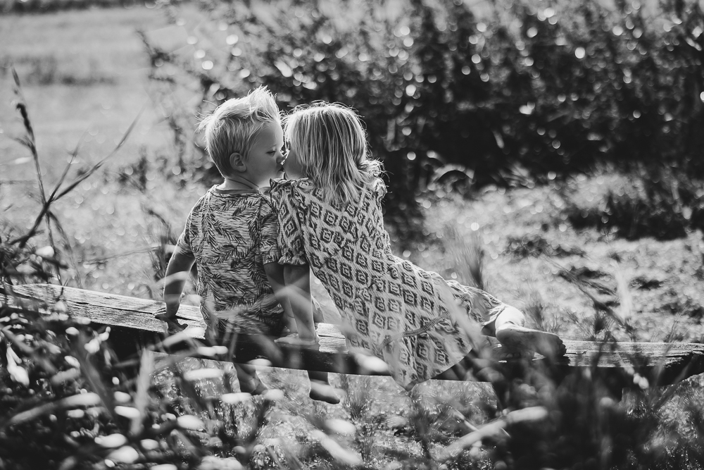 Fotograaf Friesland. Kinderfotografie. door Nickie Fotografie, in de natuur. Zus en broertje zitten op een houten bankje en geven elkaar een kusje