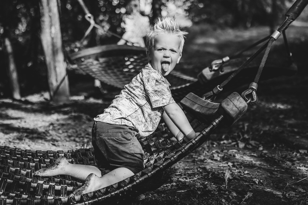 Zwart wit kinderfotografie door fotograaf NIckie Fotografie uit Friesland. Jongetje zit op een hangmat en steekst zijn tong uit.