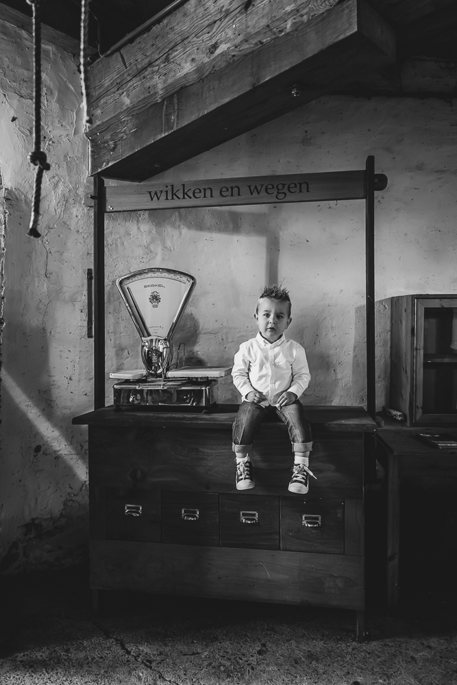 Fotograaf Friesland. Portret van peuter bij de ouderwetse weegschaal in de molen. Zwart-wit fotografie door Nickie Fotografie uit Dokkum, Friesland.