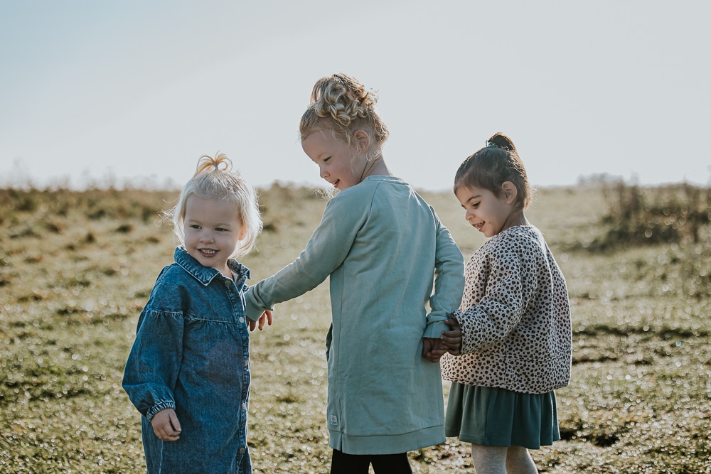 Fotoshoot Lauwersoog. Portret van 3 kleine meisjes die hand in hand lopen over het gras bij surfspot Hoek van de Bant. Lifestyle fotografie door Nickie Fotografie.