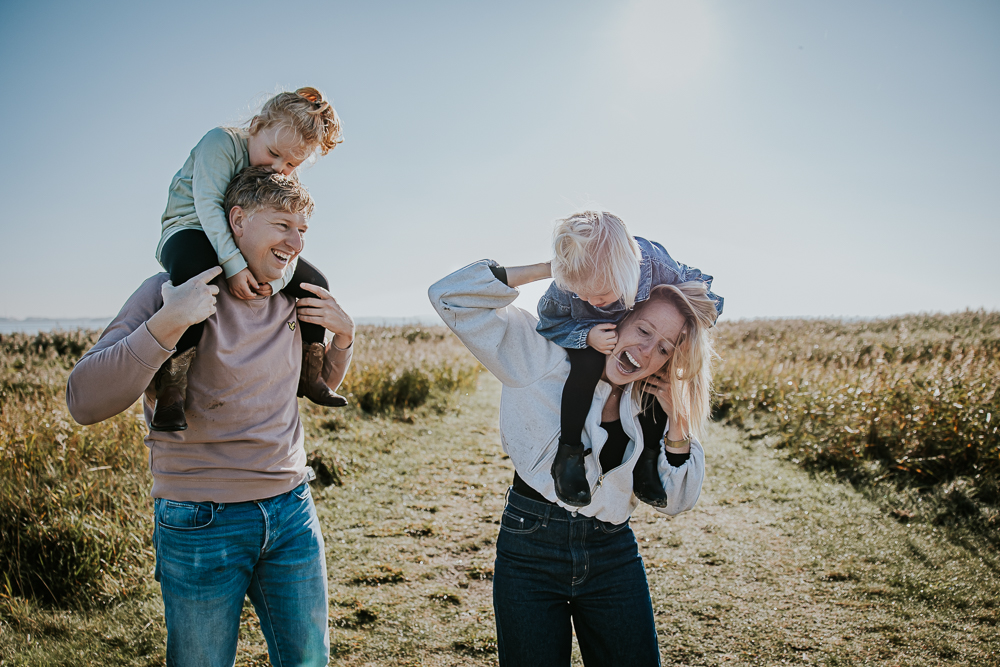 Fotoshoot Lauwersoog. Vrolijk gezinsportret door fotograaf Nickie Fotografie. De kinderen zitten bij hun ouders op de nek.