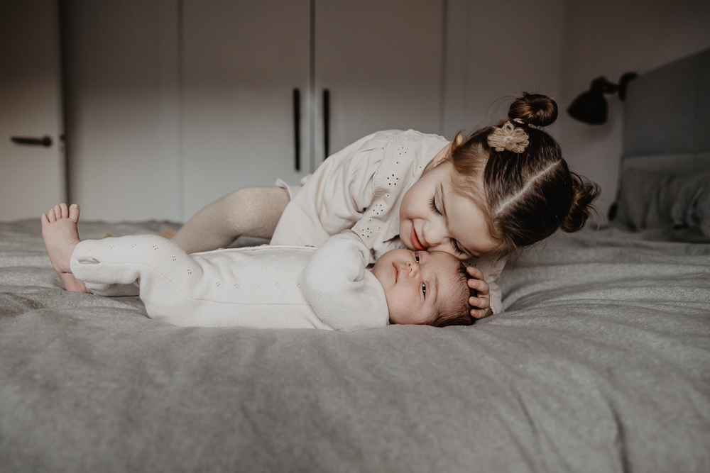 Babyshoot Leeuwarden. Grote zus knuffelt haar babyzusje op het grote bed. Fotoreportage door fotograaf Nickie Fotografie uit Friesland.