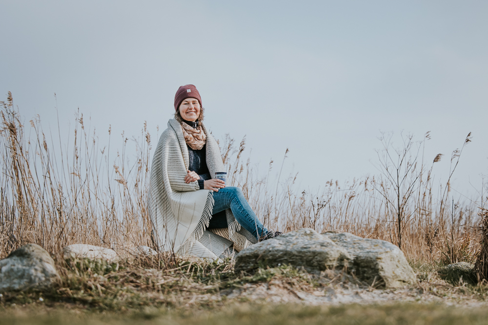 Fotoshoot in de winter. Portret van dame, gewikkeld in een warme deken en met een warme kop thee, zittend op grote keien bij het riet nabij het Lauwersmeer. Fotosessie door fotograaf Nickie Fotografie uit Dokkum, Friesland.