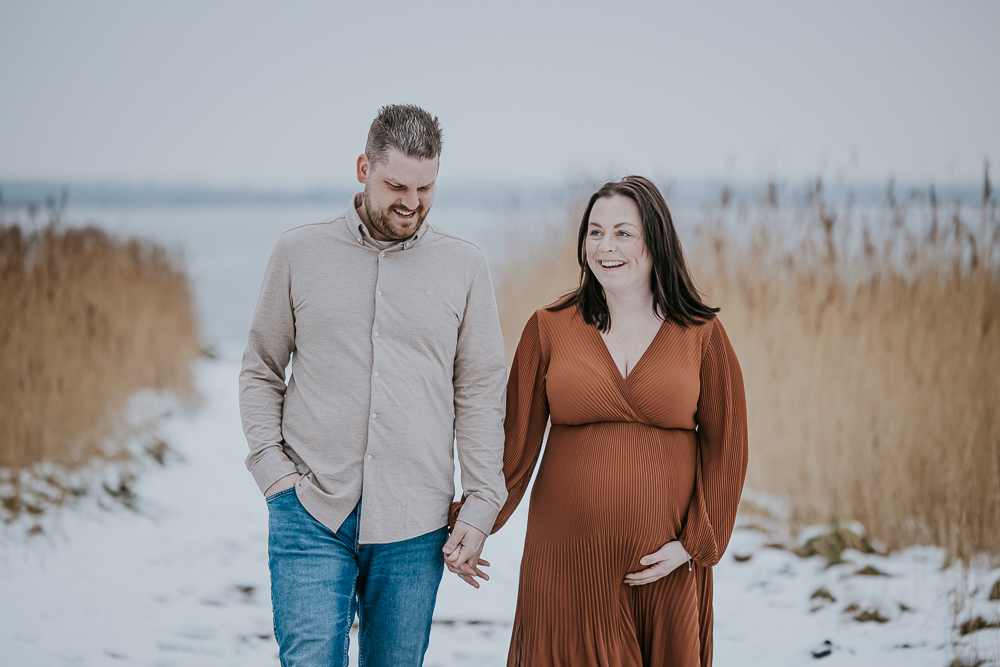 Zwangerschapsshoot met partner in de sneeuw! Inverwachting en met je partner op de foto wandelend over besneeuwde paden tussen het riet. Bolle buikshoot door zwangerschapsfotograaf Nickie Fotografie uit Dokkum, Friesland.