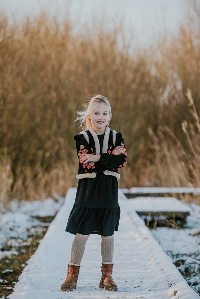 Kinderportret in de winter. Meisje staat met de armen over elkaar op de besneeuwde houten vlonder het Lauwersmeergebied. Fotoreportage door Nickie Fotografie.