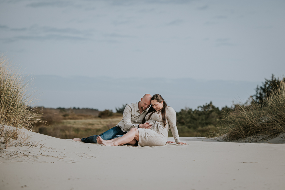 Zwangerschapsfotografie in de duinen van Ameland.  Samen in een duinpannetje voelend aan de bolle buik. Fotoreportage door fotograaf Nickie Fotografie uit Friesland.
