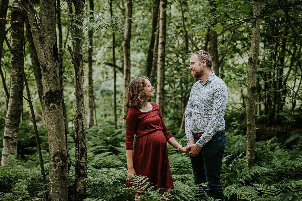 Fotograaf Friesland. Zwangerschapsshoot in De Deelen. De aanstaande ouders kijken elkaar liefdevol in de ogen aan terwijl ze tussen de varens staan. Fotoreportage door Nickie Fotografie.