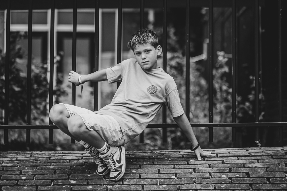 Fotoshoot Friesland. Speels portret van jongen op de hurken bij stalen hek door fotograaf Nickie Fotografie.