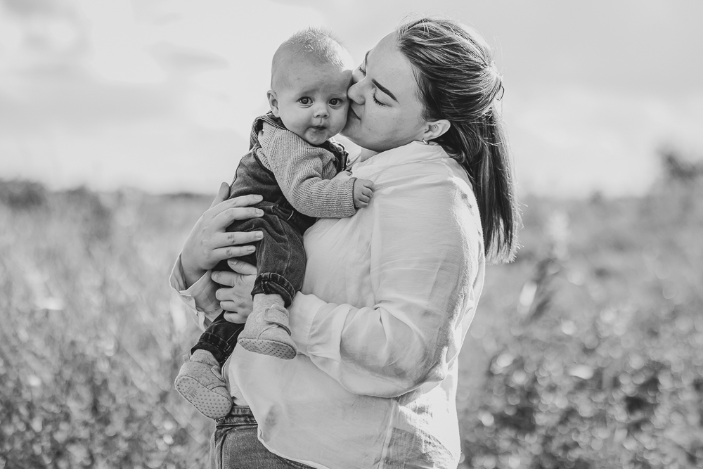 Fotoshoot Friesland. Motherhood shoot door Nickie Fotografie. Zwart-wit portret van een moeder die haar baby een kus geeft op een mooie buitenlocatie.