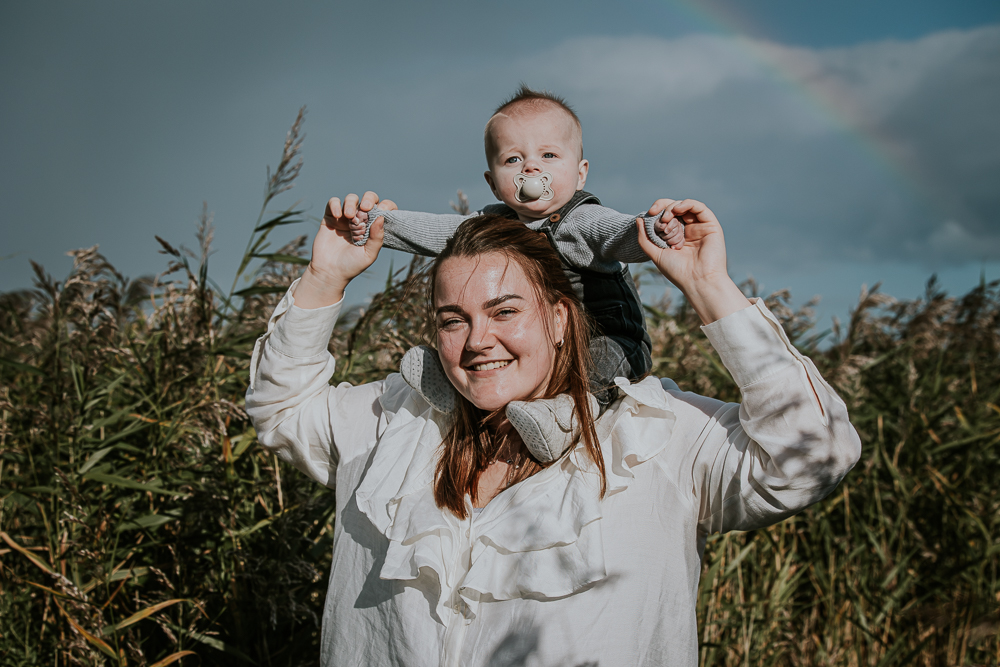 Motherhoodshoot Friesland door fotograaf Nickie Fotografie uit Dokkum. Portret van moeder en baby zoontje op haar schouders met in de achtergrond een rietkraag onder een mooie regenboog.