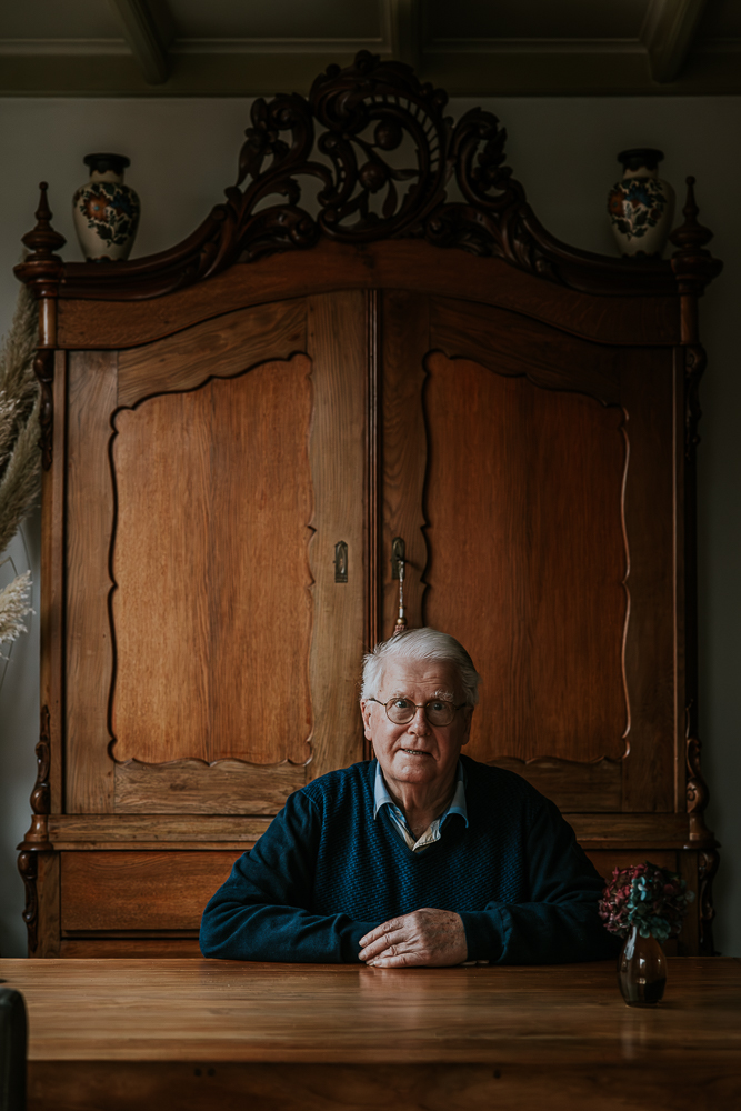 Senioren portret door fotograaf NIckie Fotografie uit Friesland. Oudere man zit thuis aan de eettafel voor een prachtige antieke kast.