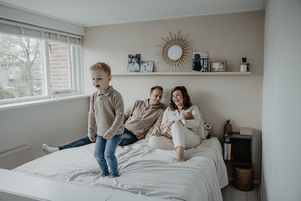 Newbornshoot Feanwâlden. Grote broer is aan het dansen op het ouderlijke bed, de ouders kijken lachend toe met de pasgeboren kleintje. Babyfotografie door newbornfotograaf Nickie Fotografie uit Friesland.