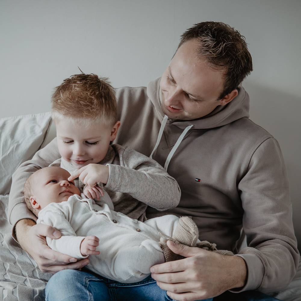 Newbornshoot Feanwâlden. Speels portret van vader met zijn twee zoontjes. Grote broer wijst naar de neus van zijn broertje. Gezinsreportage door fotograaf Nickie Fotografie uit Friesland.